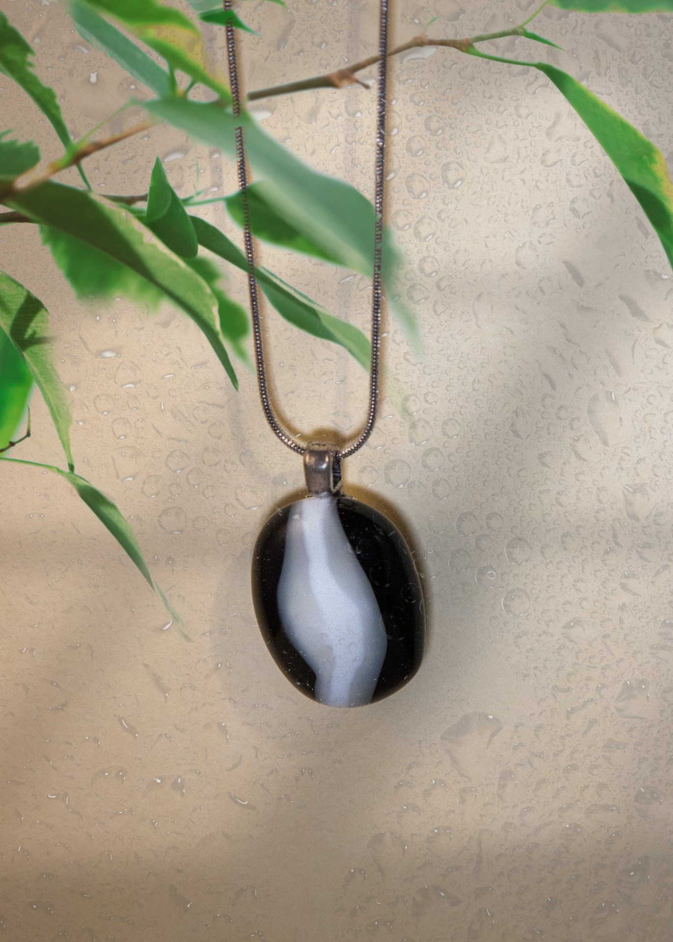 Zwarte glazen hanger aan kralenketting, hangend voor beige muur met groene bladeren en waterdruppels.