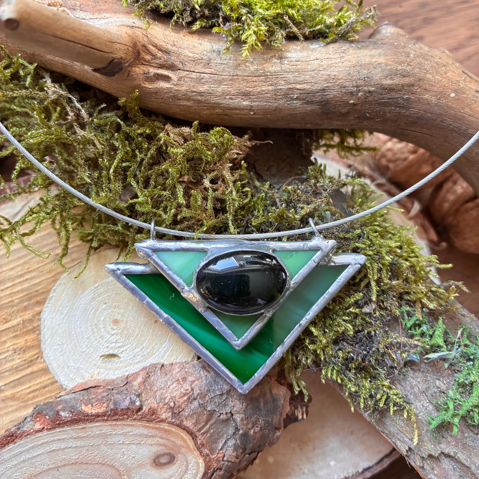 Zilveren hanger met groene driehoek en zwarte steen, op stenen onder mos en touw.